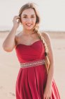 Encantadora joven con una larga coleta con elegante vestido rojo de pie en la arena y mirando a la cámara - foto de stock