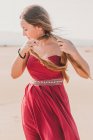 Charmante jeune femme avec une longue queue de cochon portant une robe rouge élégante debout sur le sable et regardant la caméra — Photo de stock