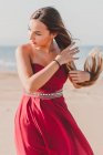Charmante junge Frau mit langem Zopf in stylischem roten Kleid steht auf Sand und blickt in die Kamera — Stockfoto