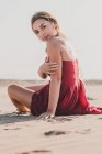 Привлекательная молодая леди с длинными светлыми волосами в стильном красном платье сидит на берегу, глядя в камеру — стоковое фото