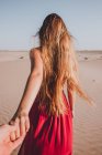 Rückansicht einer anonymen jungen Dame mit langen blonden Haaren, die ein stylisches rotes Kleid trägt und auf Sand in Richtung Kamera läuft und die Hand einer anonymen Person hält — Stockfoto