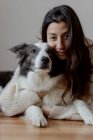 Fürsorgliche Hündin im Wollpullover umarmt lustigen Border Collie Hund, während sie gemeinsam auf dem Holzboden liegt und in die Kamera schaut — Stockfoto