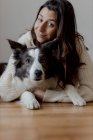 Заботливая женщина в шерстяном свитере обнимает смешную собаку Пограничного колли, лежа вместе на деревянном полу и глядя в камеру — стоковое фото