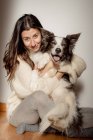 Fürsorgliche Hündin im Wollpullover umarmt lustigen Border Collie Hund, während sie gemeinsam auf dem Holzboden sitzt und in die Kamera schaut — Stockfoto
