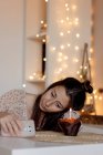 Несчастная женщина в повседневной одежде сидит за столом с вкусным кексом и читает сообщения по сотовому телефону во время празднования дня рождения в одиночестве во время карантина — стоковое фото