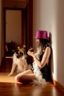 Vista lateral de sorrir feminino vestindo chapéu de festa e vestido sentado no chão com muffin e celebrando aniversário, juntamente com o cão durante a pandemia covid 19 — Fotografia de Stock