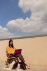 Mulher morena em roupa casual sentada na cadeira e navegando netbook roxo enquanto trabalhava e desfrutava do sol no terraço da casa durante a quarentena — Fotografia de Stock