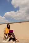 Mulher morena em roupa casual sentada na cadeira e navegando netbook roxo enquanto trabalhava e desfrutava do sol no terraço da casa durante a quarentena — Fotografia de Stock