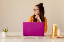 Молодая женщина в повседневной одежде и очках улыбается и смотрит на ноутбук, сидя за столом в уютной комнате — стоковое фото