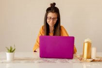 Jeune femme en tenue décontractée et lunettes souriant et regardant ordinateur portable tout en étant assis à la table dans une pièce confortable — Photo de stock