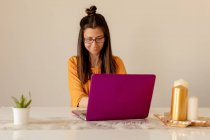 Молодая женщина в повседневной одежде и очках улыбается и смотрит на ноутбук, сидя за столом в уютной комнате — стоковое фото
