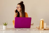 Серьезная молодая женщина в очках и повседневной одежде, работающая на розовом ноутбуке, разговаривая на смартфоне дома в карантинное время — стоковое фото