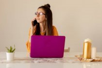 Ernste junge Frau in Brille und lässiger Kleidung arbeitet an rosa Laptop, während sie zu Hause in Quarantäne mit dem Smartphone spricht — Stockfoto
