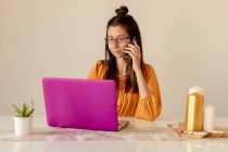 Серьезная молодая женщина в очках и повседневной одежде, работающая на розовом ноутбуке, разговаривая на смартфоне дома в карантинное время — стоковое фото