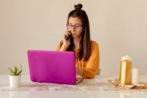 Mujer joven seria en gafas y ropa casual que trabaja en el ordenador portátil rosa mientras habla en el teléfono inteligente en casa en tiempo de cuarentena - foto de stock