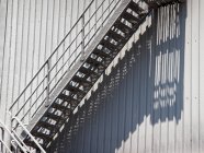 Escaleras polvorientas con barandilla colocada en la pared del edificio de metal en el día soleado dando sombra larga en la pared de tela - foto de stock