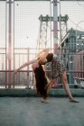 Vista lateral da jovem dançarina graciosa na saia casual realizando volta curva enquanto pé descalço na ponte na parte traseira iluminada — Fotografia de Stock