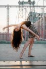 Вид збоку молодої витонченої жінки-танцюриста в повсякденній спідниці, що виконує вигин спини, стоячи босоніж на мосту в спині освітлений — стокове фото