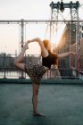 Seitenansicht einer flexiblen Frau in Freizeitkleidung, die Yoga in Tanzpose praktiziert, während sie barfuß im Gegenlicht vor dem Hintergrund der Stadtlandschaft steht und wegschaut — Stockfoto