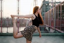 Vue latérale de la femme flexible en tenue décontractée pratiquant le yoga dans le seigneur de la pose de danse tout en se tenant pieds nus dans le dos éclairé sur fond de paysage urbain et en regardant loin — Photo de stock