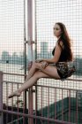 Seitenansicht der anmutigen Frau balanciert auf Metallzaun der städtischen Brücke, während sie barfuß vor dem Hintergrund der Stadtlandschaft sitzt und wegschaut — Stockfoto