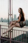 Side view of graceful woman balancing on metal fence of urban bridge while sitting barefoot on background of cityscape and looking away — Stock Photo