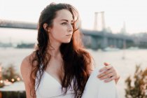 Великолепная женщина с длинными волосами в яркой одежде сидит на фоне моста и реки, расслабляясь и глядя в сторону — стоковое фото