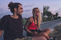 Contenu couple de hipsters itinérants assis sur le bord de la route le soir et regarder le coucher du soleil tout en se relaxant et en regardant loin — Photo de stock