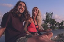 Zufriedenes Paar reisender Hipster, die abends am Straßenrand sitzen und den Sonnenuntergang beobachten, während sie sich entspannen und wegschauen — Stockfoto