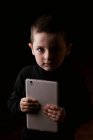 Adorable serious child in casual wear holding tablet in hands and looking at camera with determined look isolated on black background — Stock Photo
