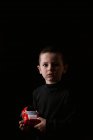 Portrait d'un petit garçon réfléchi tenant dans les mains la voiture rouge et regardant la caméra pendant la prise de vue en studio sur fond noir — Photo de stock