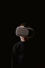 Kind trägt schwarzen Pullover und erlebt VR-Headset, während es gegen schwarze Wand steht — Stockfoto
