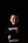 Adorable niño serio en ropa casual sosteniendo tableta en las manos y mirando a la cámara con aspecto determinado aislado sobre fondo negro - foto de stock