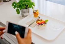 Зверху невизначена жінка фотографує зі смартфоном, сидячи за столом у ресторані та їдячи смачні здорові фруктові пустелі на білій тарілці — стокове фото