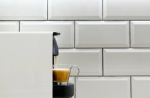 Caffettiera moderna cialde versando espresso caldo in tazza di vetro su sfondo di piastrelle di ceramica sulla parete della cucina — Foto stock