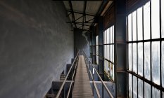 Schmale Holzbrücke mit Stahlgeländern über Räumen mit grauen Betonwänden und staubigen, schmutzigen, großen Fenstern in verlassenen Fabriken — Stockfoto