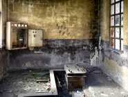Destroyed dirty room with layer of dust and pieces of concrete on floor covered of moss broken window and old inoperative transformer hanging on shabby wall with black spots in abandoned building — Stock Photo