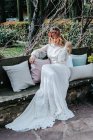 Elegante junge Frau in weißem Kleid und Blumenkranz sitzt am Hochzeitstag im Garten inmitten von Kissen auf einer Bank — Stockfoto