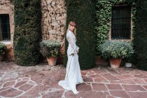 Seitenansicht einer jungen Frau in elegantem weißen Kleid und Blumenkranz, die in die Kamera schaut, während sie in der Nähe eines mit Weinreben und Topfsträuchern dekorierten Hauses am Hochzeitstag steht — Stockfoto