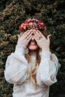 Задоволена молода жінка в весільному вбранні і квітковому вінку посміхається і прикриває очі, стоячи біля зеленого чагарника в саду — стокове фото