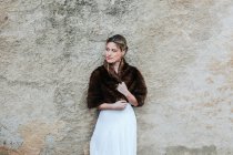 Young woman in white dress and short fur coat looking away while standing near weathered building wall on wedding day — Stock Photo