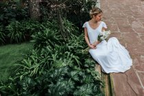 From above elegant young woman in white dress and with bouquet sitting on shabby path near bushes and looking away on wedding day in garden — Stock Photo