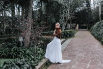 Side view of young lady in white dress and short fur coat looking at camera while stepping on border of weathered path during wedding in green garden — Stock Photo