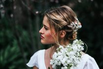 Гарненька блондинка з купою білих квітів, дивлячись далеко, стоячи на розмитому фоні саду під час весілля в літній день — стокове фото