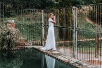 Вид збоку молодої жінки в білій весільній сукні, що стоїть на обвітрюваній межі біля металевого паркану і спокійного басейну в саду — стокове фото