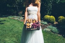 Unrecognizable young bride in elegant white wedding dress standing on green lawn in garden and holding basket with fresh buns and bread — Stock Photo