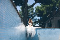 Снизу вид молодой мечтательной женщины в элегантном белом свадебном платье, отдыхающей на краю пустого бассейна возле зеленого дерева и мечтательно смотрящей в сторону — стоковое фото
