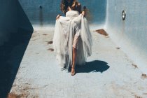 Щаслива молода анонімна наречена в елегантній білій весільній сукні чуттєво танцює в порожньому брудному басейні в сонячний день під час романтичної вечірки — стокове фото