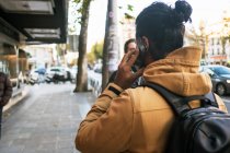 Vista trasera de hombre anónimo en chaqueta de otoño con pelo largo y oscuro en bollo hablando en smartphone mientras camina por la calle en la ciudad con mochila - foto de stock