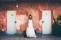 Charming young bride in elegant white wedding dress standing near red shabby wall near toilet doors and looking at camera — Stock Photo